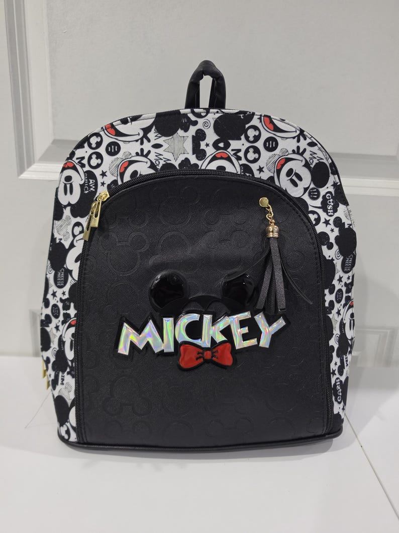 May include: A black and white backpack featuring a repeating Mickey Mouse pattern on the sides and top. The front pocket is solid black with a debossed Mickey icon pattern, a holographic MICKEY text logo, and a red bow tie accent.