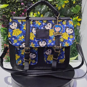 Puede incluir: Un bolso tipo cartera negro con un panel de tela azul que presenta un patr&oacute;n repetitivo de personajes de Coraline y flores amarillas. El bolso tiene un asa superior, una correa para el hombro, dos bolsillos frontales con hebillas doradas y un dije de Coraline.