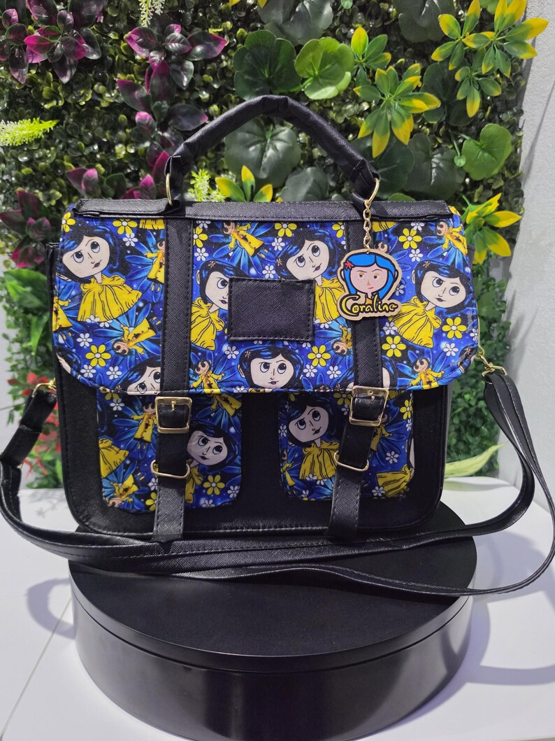 Puede incluir: Un bolso tipo cartera negro con un panel de tela azul que presenta un patr&oacute;n repetitivo de personajes de Coraline y flores amarillas. El bolso tiene un asa superior, una correa para el hombro, dos bolsillos frontales con hebillas doradas y un dije de Coraline.
