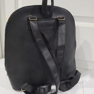 May include: A black backpack featuring a smooth, solid-colored back panel, two adjustable shoulder straps, and a top carry handle. The bag includes gold-toned metal hardware attachments for the straps and a zipper closure visible along the side edge.