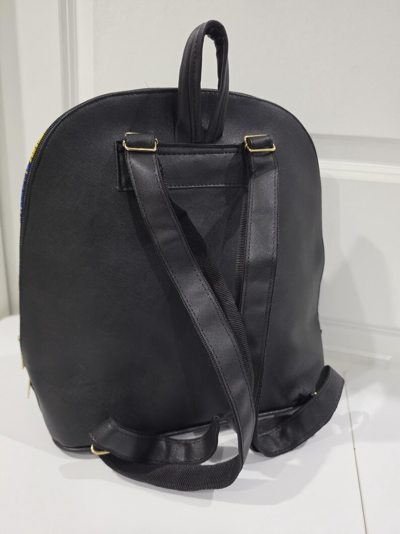 May include: A black backpack featuring a smooth, solid-colored back panel, two adjustable shoulder straps, and a top carry handle. The bag includes gold-toned metal hardware attachments for the straps and a zipper closure visible along the side edge.