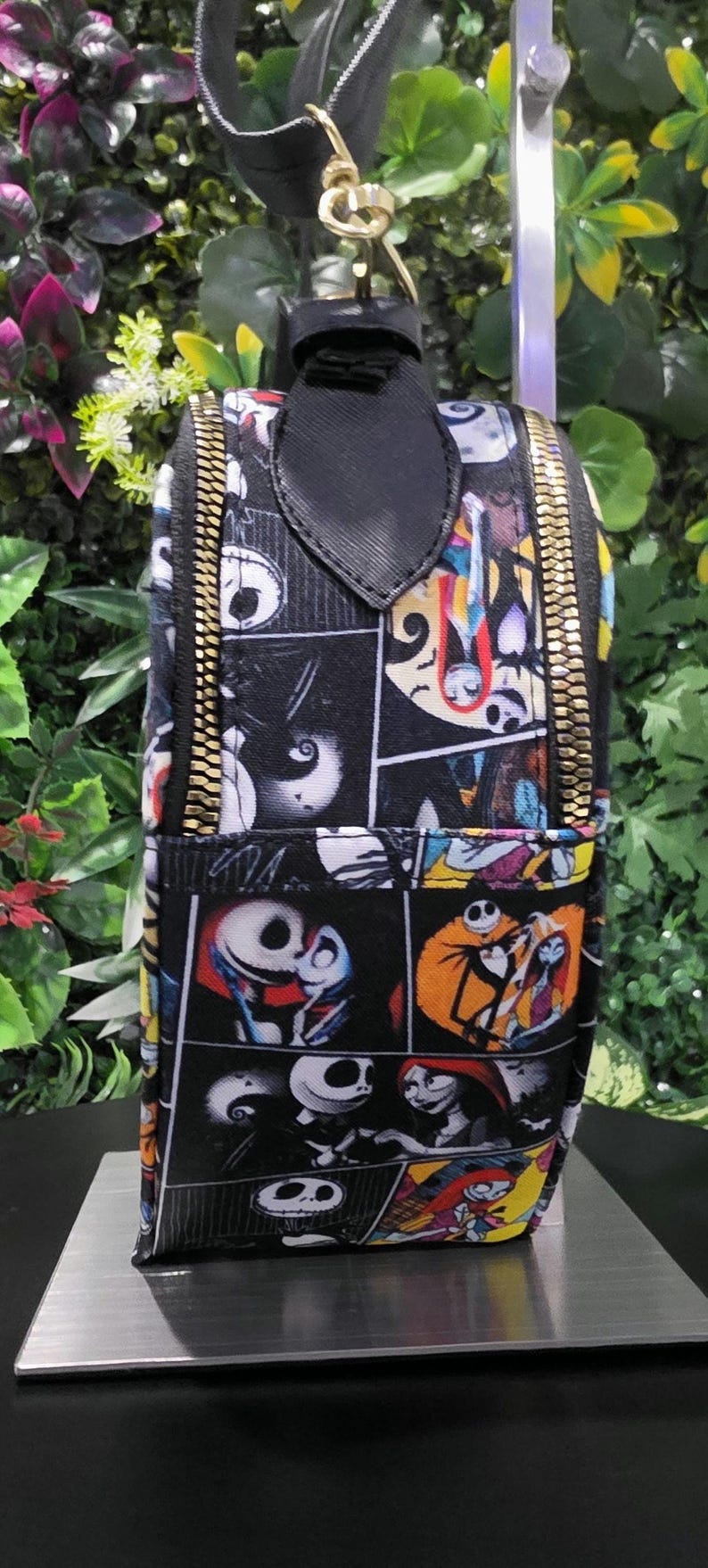 May include: A rectangular mini backpack featuring a comic book style print of Jack Skellington and Sally from The Nightmare Before Christmas. The bag has a black top handle, gold-toned metal zippers, and a black strap with a gold-toned metal clasp.