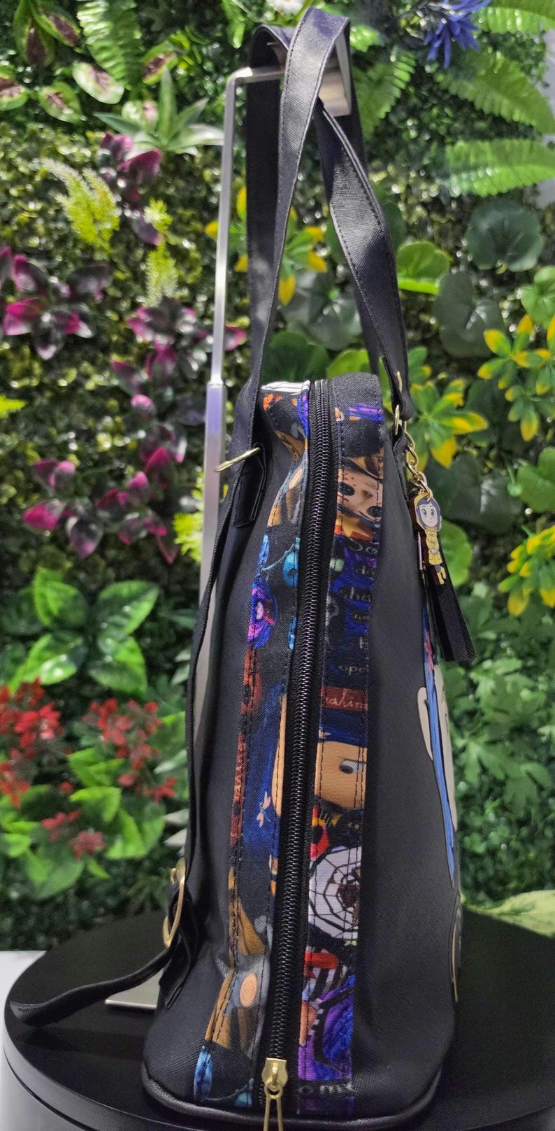 Puede incluir: Una mochila negra que presenta un panel lateral con un estampado de personajes coloridos, un cierre de cremallera negro y un peque&ntilde;o dije colgante unido al asa superior. La bolsa tiene correas negras ajustables y un dise&ntilde;o vertical.