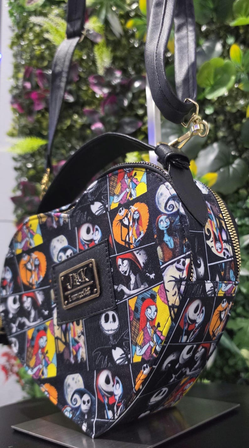 May include: A heart-shaped crossbody bag featuring a black and white grid pattern with colorful illustrations of Jack Skellington and Sally from The Nightmare Before Christmas. The bag has a black top handle, a shoulder strap, and a gold-tone metal plaque.