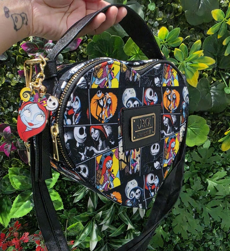 May include: A heart-shaped crossbody bag featuring a black, yellow, and white comic-style print of Jack Skellington and Sally. The bag includes a black top handle, a gold-tone zipper, and a metal plaque with the text JACK LOUNGEFLY.