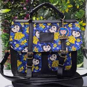 Puede incluir: Un bolso tipo cartera negro con un panel de tela azul que presenta un patr&oacute;n repetitivo de un personaje de dibujos animados con cabello azul y chubasquero amarillo, decorado con flores amarillas. Incluye dos bolsillos y un dije de Coraline.