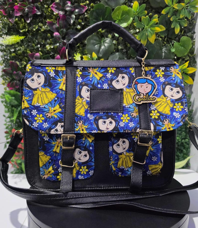 Puede incluir: Un bolso tipo cartera negro con un panel de tela azul que presenta un patr&oacute;n repetitivo de un personaje de dibujos animados con cabello azul y chubasquero amarillo, decorado con flores amarillas. Incluye dos bolsillos y un dije de Coraline.
