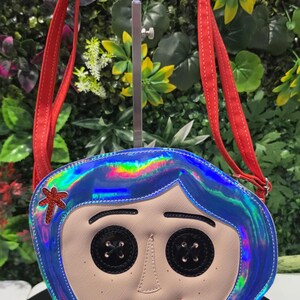 May include: A crossbody bag shaped like a character face with iridescent blue hair, black button eyes, and a red hair clip. The bag features a light beige face, a small red mouth, and two red fabric shoulder straps attached to the top edges.