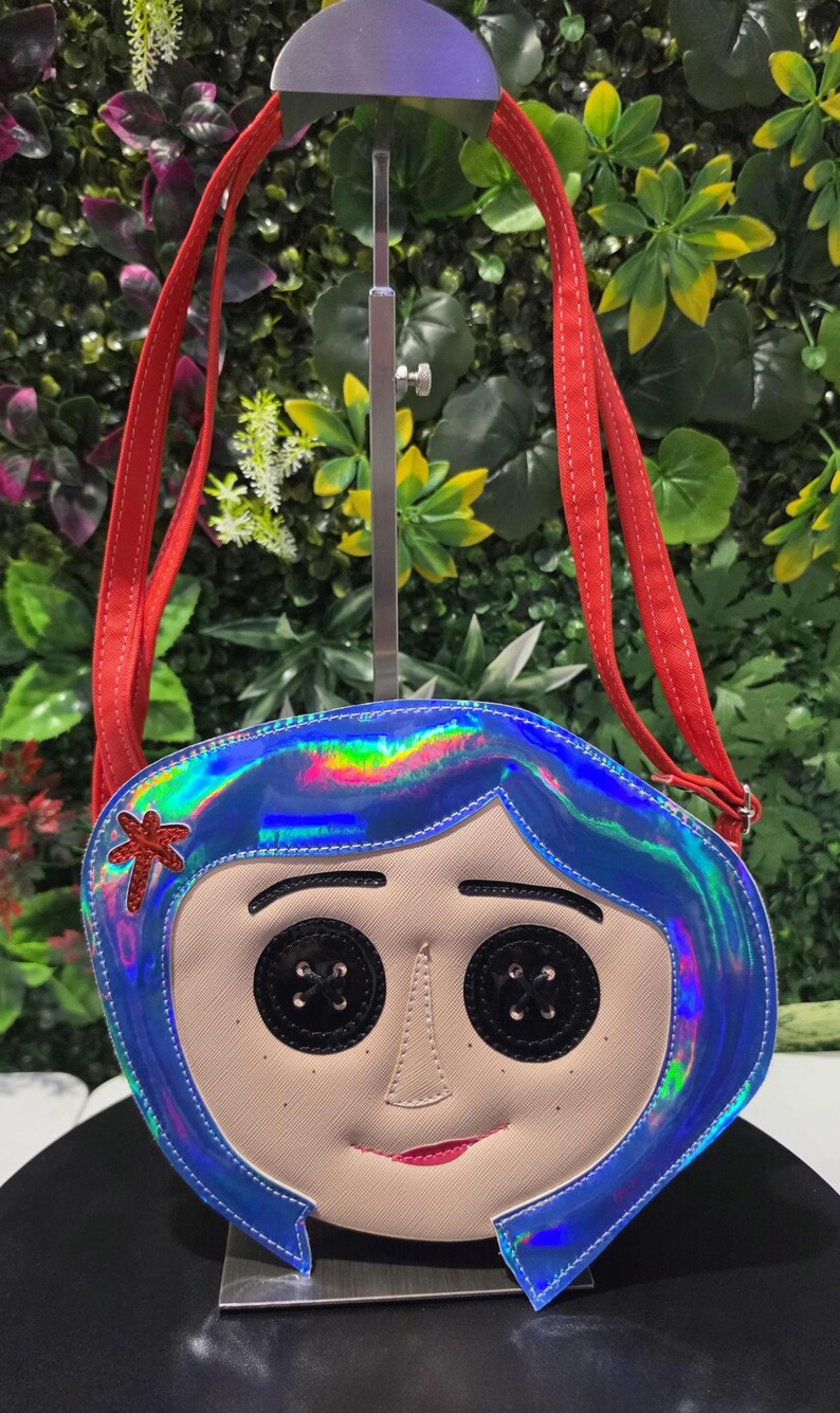 May include: A crossbody bag shaped like a character face with iridescent blue hair, black button eyes, and a red hair clip. The bag features a light beige face, a small red mouth, and two red fabric shoulder straps attached to the top edges.