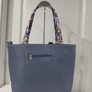 May include: A slate blue tote bag with a smooth, leather-like texture. The bag features a horizontal zippered pocket on the front with a gold-toned pull. Two shoulder straps are attached, decorated with a colorful, repeating cartoon character pattern.