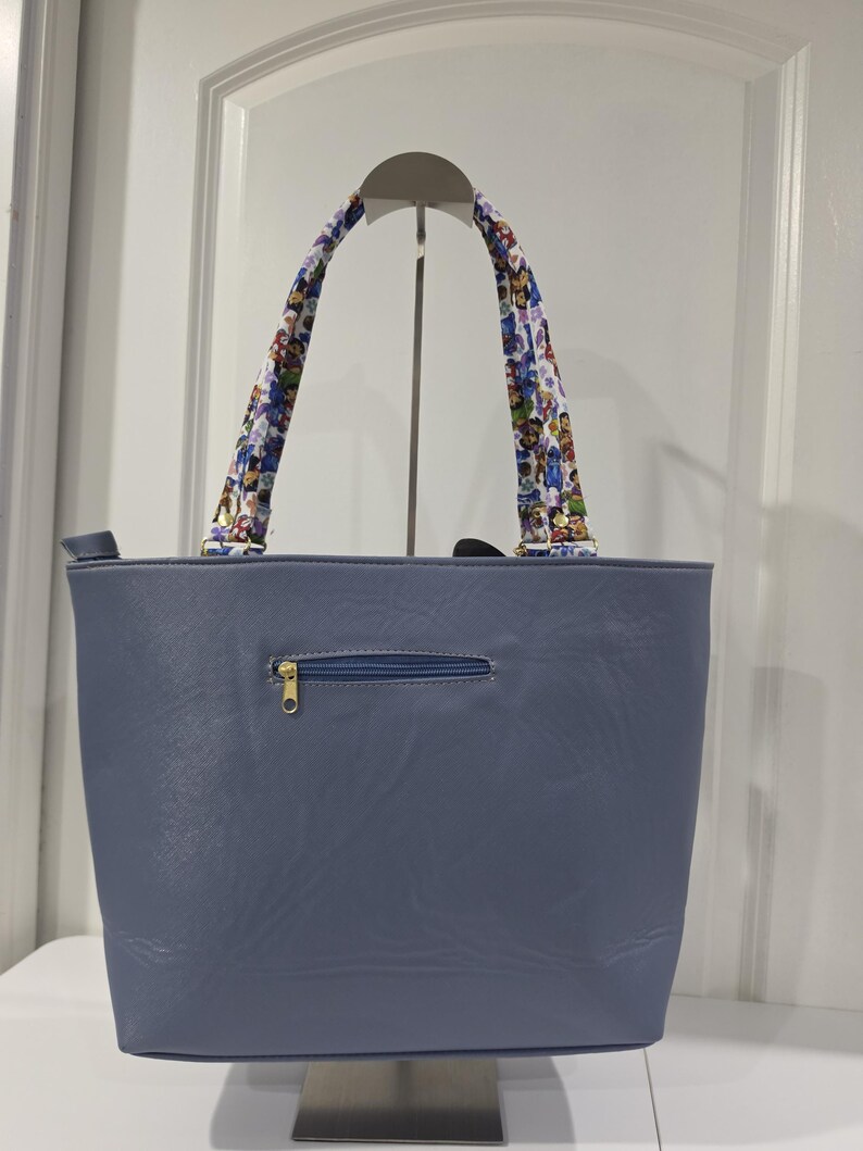 May include: A slate blue tote bag with a smooth, leather-like texture. The bag features a horizontal zippered pocket on the front with a gold-toned pull. Two shoulder straps are attached, decorated with a colorful, repeating cartoon character pattern.
