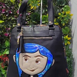 Puede incluir: Un bolso tote negro con un rostro grande en relieve, cabello azul iridiscente y el nombre Coraline en letras doradas. El bolso incluye dos asas superiores, un bolsillo frontal con cremallera y un peque&ntilde;o dije colgante del personaje.