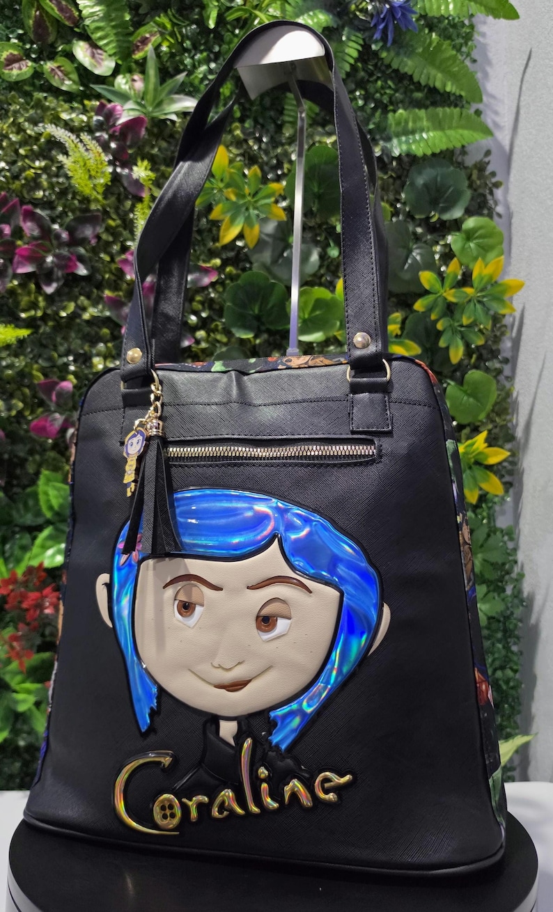 Puede incluir: Un bolso tote negro con un rostro grande en relieve, cabello azul iridiscente y el nombre Coraline en letras doradas. El bolso incluye dos asas superiores, un bolsillo frontal con cremallera y un peque&ntilde;o dije colgante del personaje.