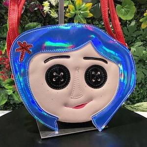 May include: A crossbody bag shaped like a face with iridescent blue hair, black button eyes, a small red hair clip, and a light beige complexion. The bag features red shoulder straps and a subtle smile, capturing a stylized character design.
