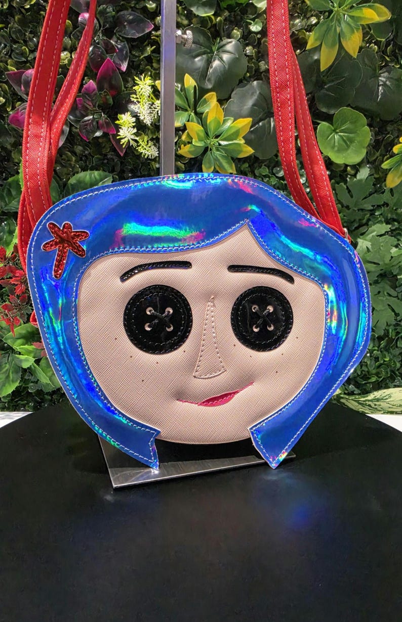 May include: A crossbody bag shaped like a face with iridescent blue hair, black button eyes, a small red hair clip, and a light beige complexion. The bag features red shoulder straps and a subtle smile, capturing a stylized character design.