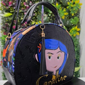 May include: A black dome-shaped handbag featuring a prominent graphic of a character with blue hair and a star hair clip. The bag has a textured surface with embossed patterns, a black tassel, and the word Coraline in gold lettering on the front.