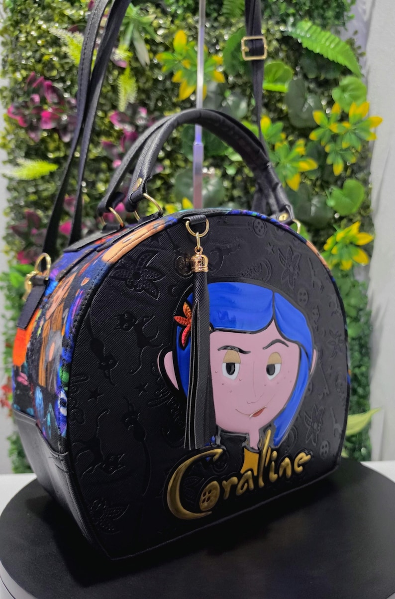 May include: A black dome-shaped handbag featuring a prominent graphic of a character with blue hair and a star hair clip. The bag has a textured surface with embossed patterns, a black tassel, and the word Coraline in gold lettering on the front.