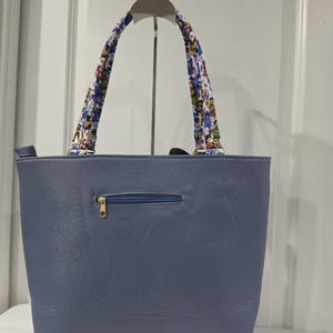 May include: A slate blue tote bag with a smooth, leather-like texture, featuring a horizontal zippered pocket on the front. The bag has two shoulder straps decorated with a colorful, repeating cartoon character pattern and gold-toned hardware.