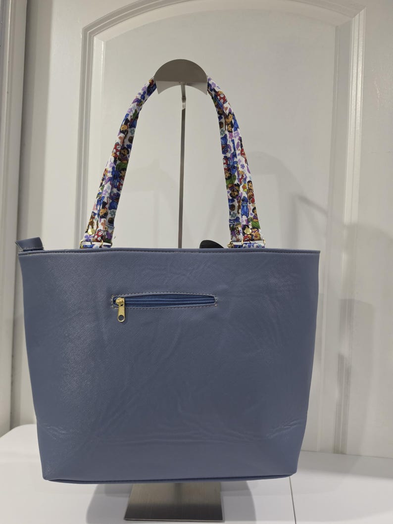 May include: A slate blue tote bag with a smooth, leather-like texture, featuring a horizontal zippered pocket on the front. The bag has two shoulder straps decorated with a colorful, repeating cartoon character pattern and gold-toned hardware.