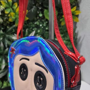 May include: A round crossbody bag featuring a character face with black button eyes, a small red mouth, and blue iridescent hair. The bag has a black textured side panel, a red zipper closure, and two red adjustable shoulder straps with white stitching.