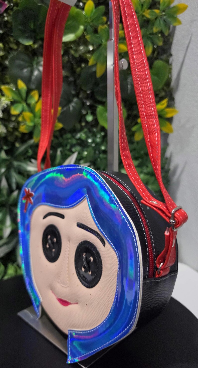 May include: A round crossbody bag featuring a character face with black button eyes, a small red mouth, and blue iridescent hair. The bag has a black textured side panel, a red zipper closure, and two red adjustable shoulder straps with white stitching.