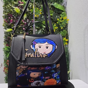 以下が含まれることがあります： フロントフラップにコララインのキャラクターアップリケと金色の文字でCoralineと書かれた黒い構造のハンドバッグ。下部にはコララインのキャラクターと黒猫のカラフルなグラフィックプリントが施され、タッセルのアクセントが付いています。