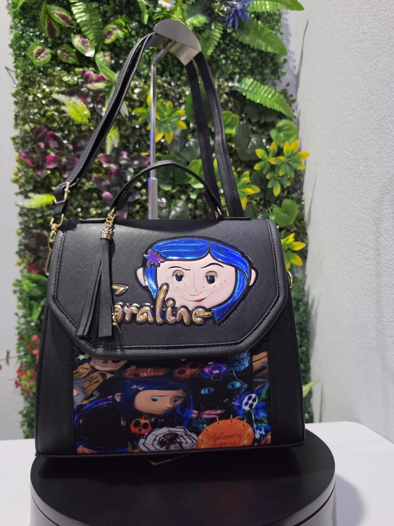 以下が含まれることがあります： フロントフラップにコララインのキャラクターアップリケと金色の文字でCoralineと書かれた黒い構造のハンドバッグ。下部にはコララインのキャラクターと黒猫のカラフルなグラフィックプリントが施され、タッセルのアクセントが付いています。