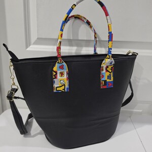 Pu&ograve; includere: Una borsa tote nera con due manici caratterizzati da un motivo colorato a quadrati rossi, gialli, blu e bianchi con sagome di personaggi dei cartoni animati e la scritta "OH BOY". La borsa include una tracolla nera rimovibile con dettagli dorati.
