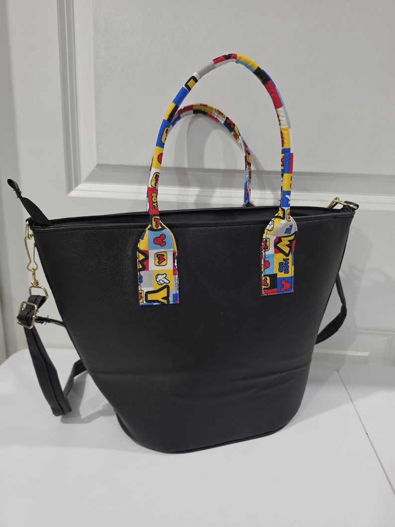 Pu&ograve; includere: Una borsa tote nera con due manici caratterizzati da un motivo colorato a quadrati rossi, gialli, blu e bianchi con sagome di personaggi dei cartoni animati e la scritta "OH BOY". La borsa include una tracolla nera rimovibile con dettagli dorati.