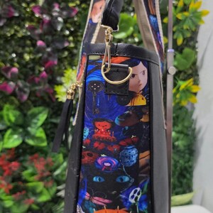 May include: A black tote bag featuring a colorful patterned side panel with illustrations of characters, a black cat with blue eyes, and an orange pumpkin with the text Welcome home! The bag has a gold-tone metal D-ring and a wide patterned shoulder strap.