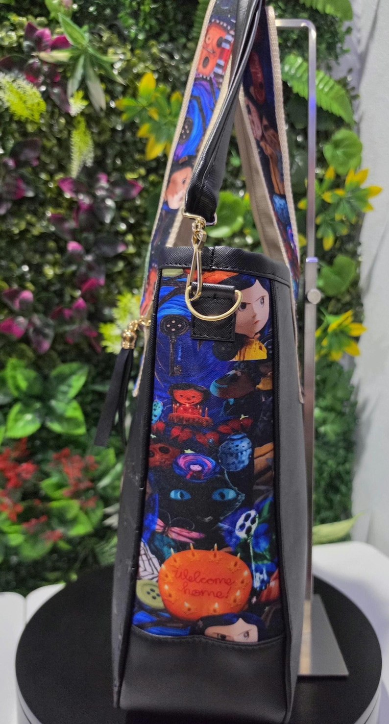 May include: A black tote bag featuring a colorful patterned side panel with illustrations of characters, a black cat with blue eyes, and an orange pumpkin with the text Welcome home! The bag has a gold-tone metal D-ring and a wide patterned shoulder strap.