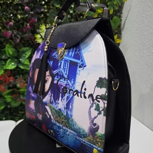 May include: A black structured handbag featuring a printed graphic of the character Coraline and a house on the front panel. The bag has a top handle, a gold-toned metal clasp closure, and a black tassel charm attached to a gold chain.