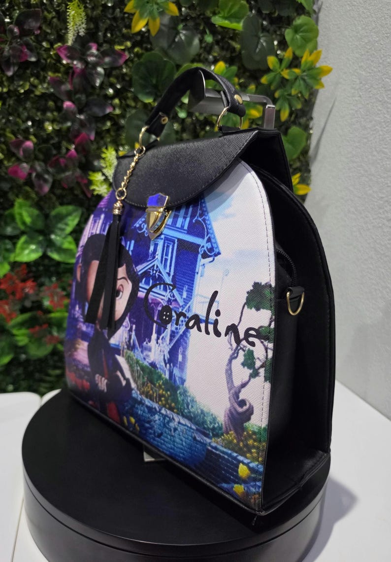 May include: A black structured handbag featuring a printed graphic of the character Coraline and a house on the front panel. The bag has a top handle, a gold-toned metal clasp closure, and a black tassel charm attached to a gold chain.
