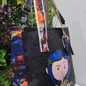 May include: A black tote bag featuring an embossed pattern of keys and buttons. The side panel and straps display a colorful Coraline-themed print. A large, raised Coraline character applique with blue hair is attached to the front with the name Coraline.