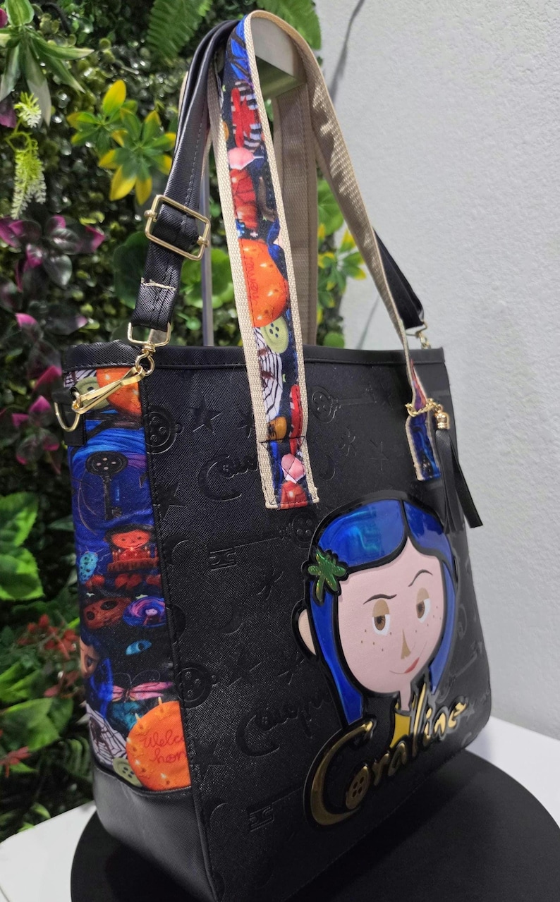May include: A black tote bag featuring an embossed pattern of keys and buttons. The side panel and straps display a colorful Coraline-themed print. A large, raised Coraline character applique with blue hair is attached to the front with the name Coraline.