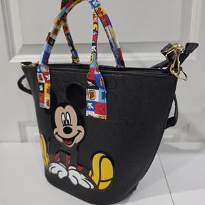 Pu&ograve; includere: Una borsa tote nera con un motivo in rilievo di Mickey Mouse sulla superficie e una grande grafica colorata di Mickey Mouse sul davanti. La borsa include due manici con un motivo multicolore composto da quadrati rossi, blu e gialli.