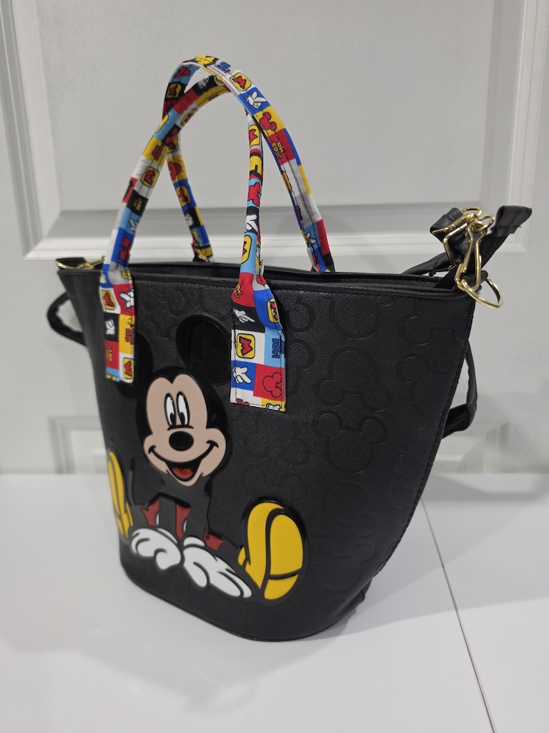Pu&ograve; includere: Una borsa tote nera con un motivo in rilievo di Mickey Mouse sulla superficie e una grande grafica colorata di Mickey Mouse sul davanti. La borsa include due manici con un motivo multicolore composto da quadrati rossi, blu e gialli.