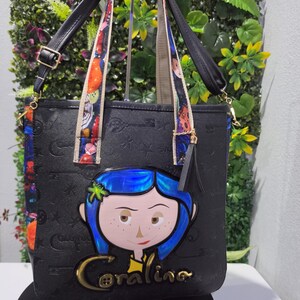 May include: A black tote bag featuring an embossed pattern of keys and stars, with a central graphic of a blue-haired character and the text Coraline. The bag includes black handles, colorful patterned straps, and a black tassel charm on the side.
