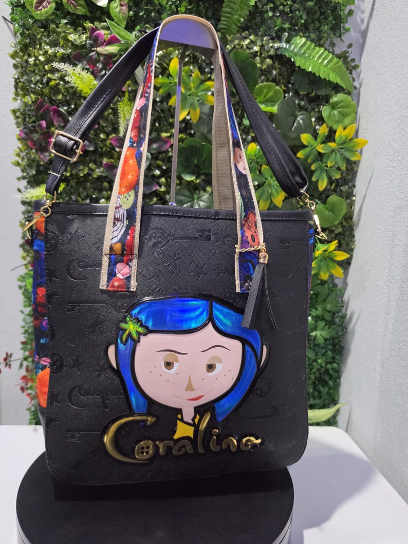 May include: A black tote bag featuring an embossed pattern of keys and stars, with a central graphic of a blue-haired character and the text Coraline. The bag includes black handles, colorful patterned straps, and a black tassel charm on the side.