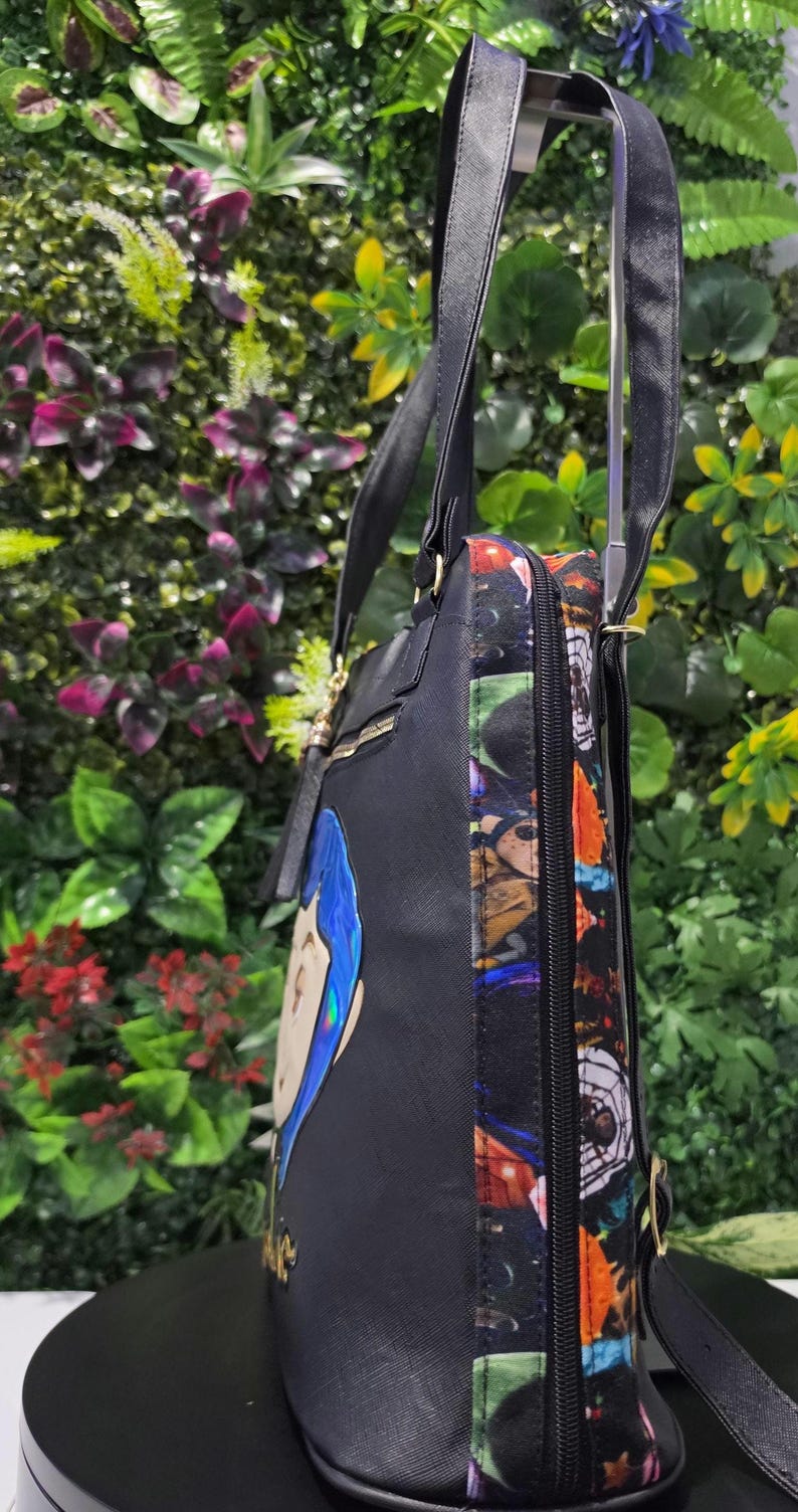 Puede incluir: Un bolso tote negro con dos correas para el hombro y un panel lateral con un estampado gr&aacute;fico colorido. La parte frontal del bolso muestra una ilustraci&oacute;n de personaje estilizada con cabello azul y letras doradas. Cierre de cremallera lateral.
