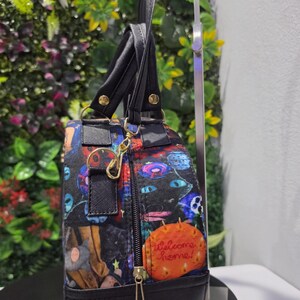 May include: A black handbag with a colorful graphic print featuring various characters and an orange circular patch with the text Welcome home! in cursive. The bag has black handles, a gold-toned metal clasp, and a black zipper with a gold pull.