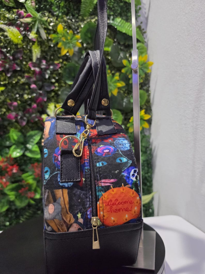May include: A black handbag with a colorful graphic print featuring various characters and an orange circular patch with the text Welcome home! in cursive. The bag has black handles, a gold-toned metal clasp, and a black zipper with a gold pull.