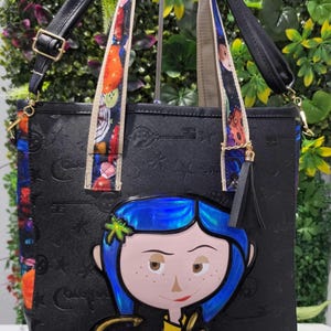 May include: A black tote bag featuring an embossed pattern of keys and stars. The front displays a large, colorful character applique with blue hair and the name Coraline in gold lettering. It includes patterned shoulder straps and a black tassel.