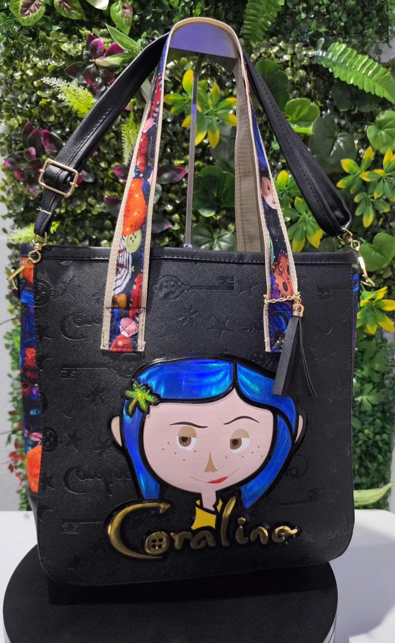 May include: A black tote bag featuring an embossed pattern of keys and stars. The front displays a large, colorful character applique with blue hair and the name Coraline in gold lettering. It includes patterned shoulder straps and a black tassel.