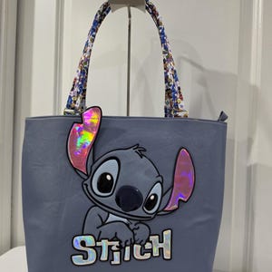 May include: A light blue tote bag featuring a large graphic of the character Stitch with iridescent pink ears and the name Stitch in iridescent lettering. The bag has two shoulder straps with a colorful floral pattern and a zipper closure.