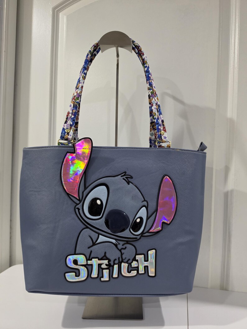 May include: A light blue tote bag featuring a large graphic of the character Stitch with iridescent pink ears and the name Stitch in iridescent lettering. The bag has two shoulder straps with a colorful floral pattern and a zipper closure.