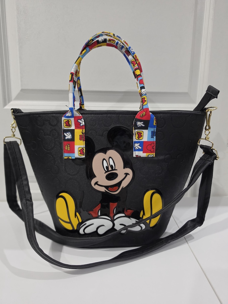 Pu&ograve; includere: Una borsa tote nera con un motivo di Topolino in rilievo all'esterno e una grande grafica colorata di Topolino sul davanti. La borsa include due manici superiori con un motivo multicolore e una tracolla nera rimovibile.