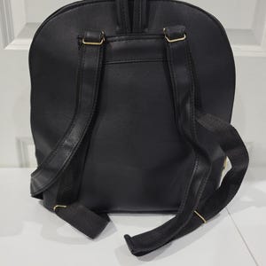 May include: A black backpack featuring a textured surface, a top handle, and two adjustable shoulder straps. The bag includes gold-toned metal hardware connecting the straps to the main body, providing a secure and functional design for carrying items.