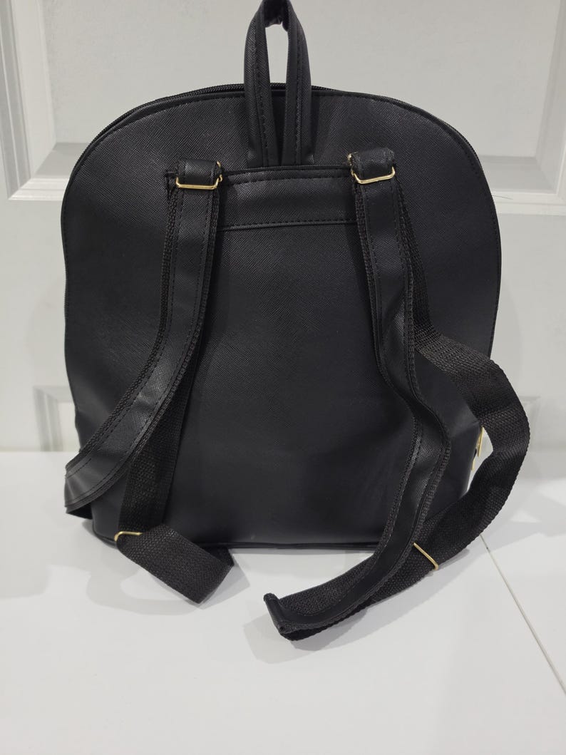 May include: A black backpack featuring a textured surface, a top handle, and two adjustable shoulder straps. The bag includes gold-toned metal hardware connecting the straps to the main body, providing a secure and functional design for carrying items.