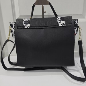 May include: A black handbag featuring a top handle and a detachable shoulder strap. The flap and side panels are decorated with a black and white pattern of Mickey Mouse faces and the text CHEESE. The main body of the bag is solid black.